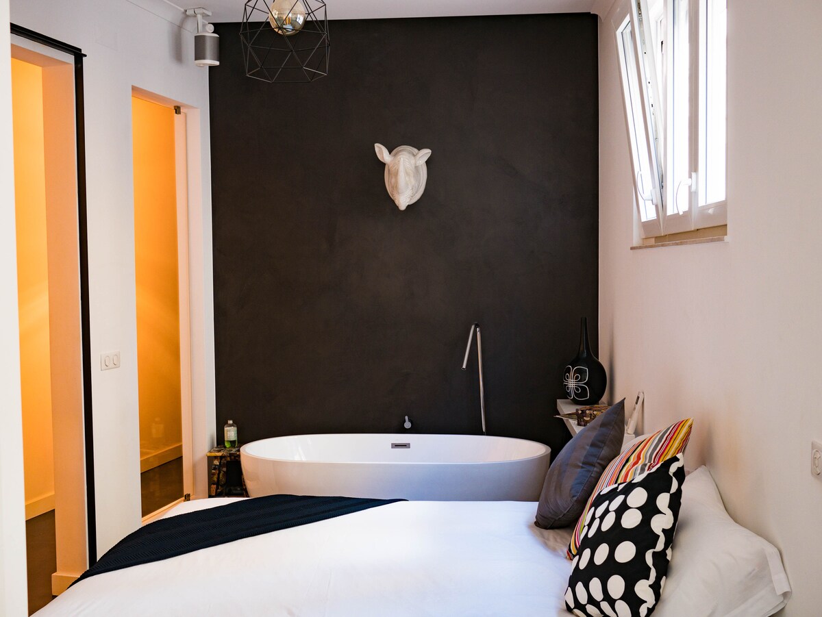 A spacious bedroom features a minimalist design, highlighted by a large freestanding bathtub and a dark accent wall. Decorative elements include a mounted wall art piece and a bedside table with a small lamp. Natural light enters through a nearby window, creating a serene atmosphere.