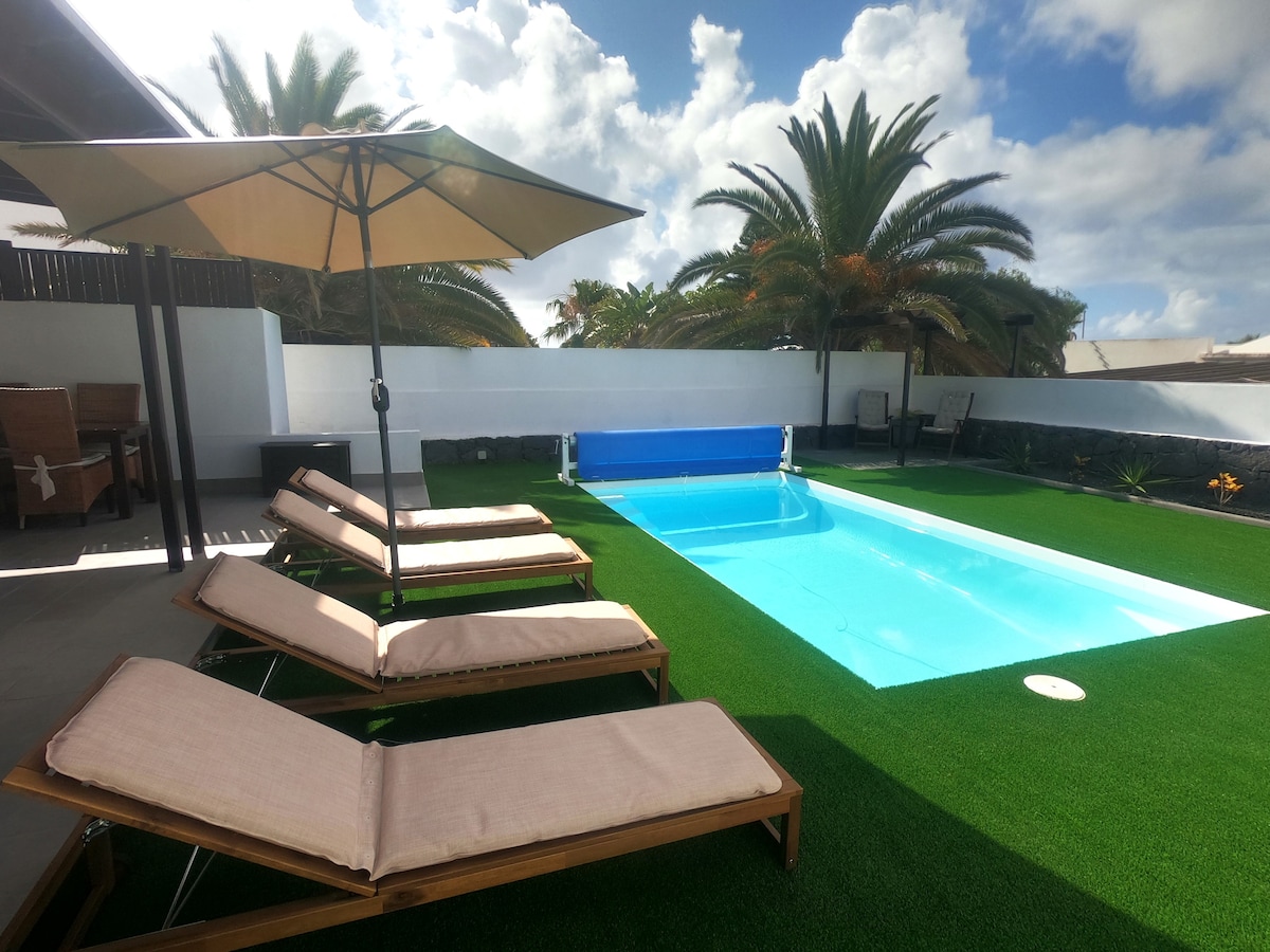 A private heated pool, measuring 6 x 3 meters, is surrounded by a green lawn. Lounge chairs with cushioned seats are positioned under an umbrella for shade. Palm trees are visible in the background, contributing to a relaxed outdoor environment.