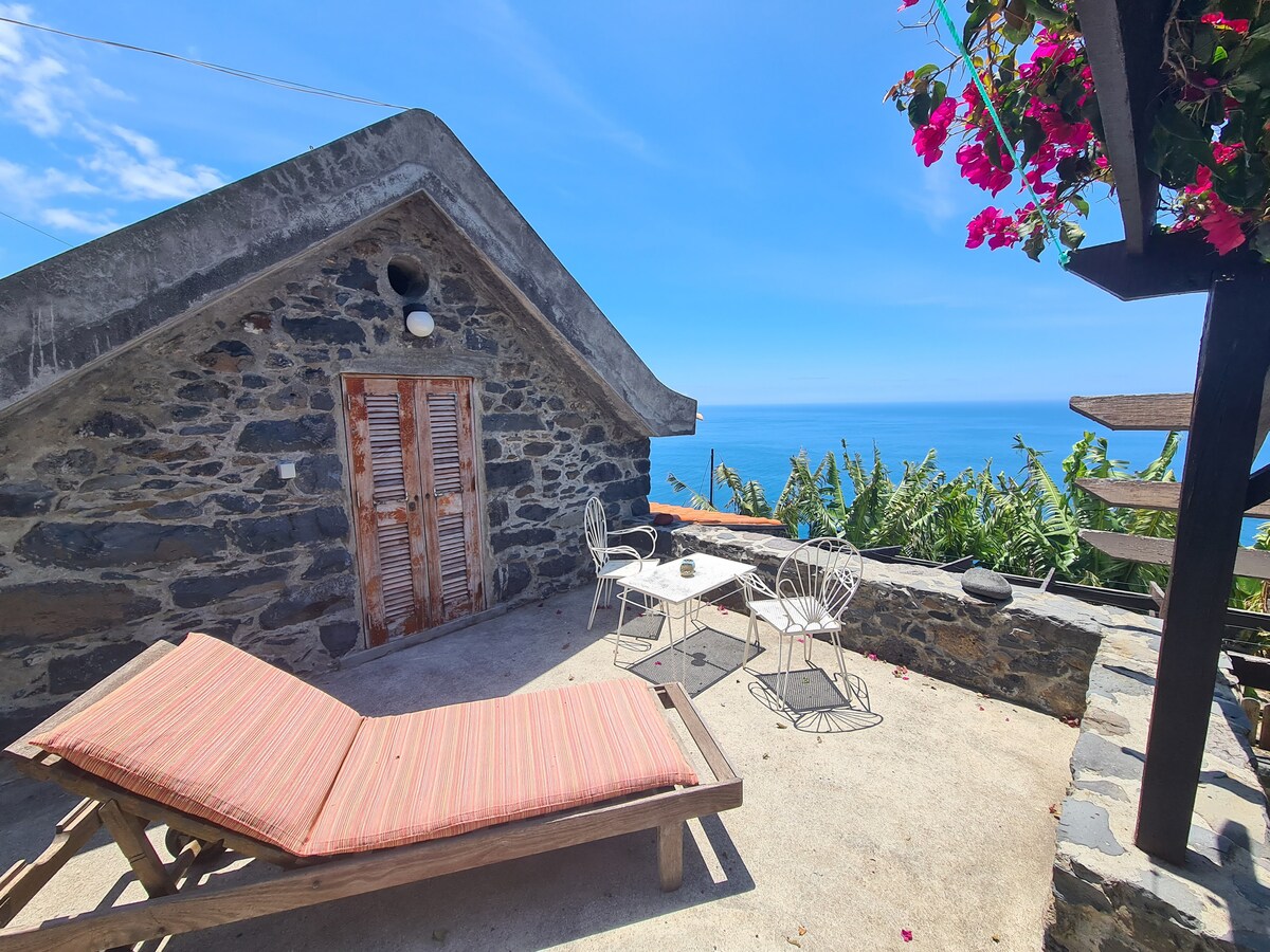 A stone terrace features a sun lounger and a small, round table surrounded by four chairs. A spiral staircase leads to a higher level. The ocean vista is visible in the background, with a clear blue sky overhead and flowering plants adding a touch of color.