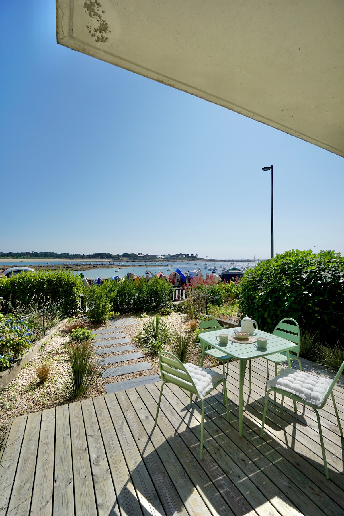 A wooden terrace displays a small round table and two green chairs, set against a backdrop of lush greenery. The view features a serene waterfront landscape under clear blue skies, inviting relaxation and outdoor enjoyment.