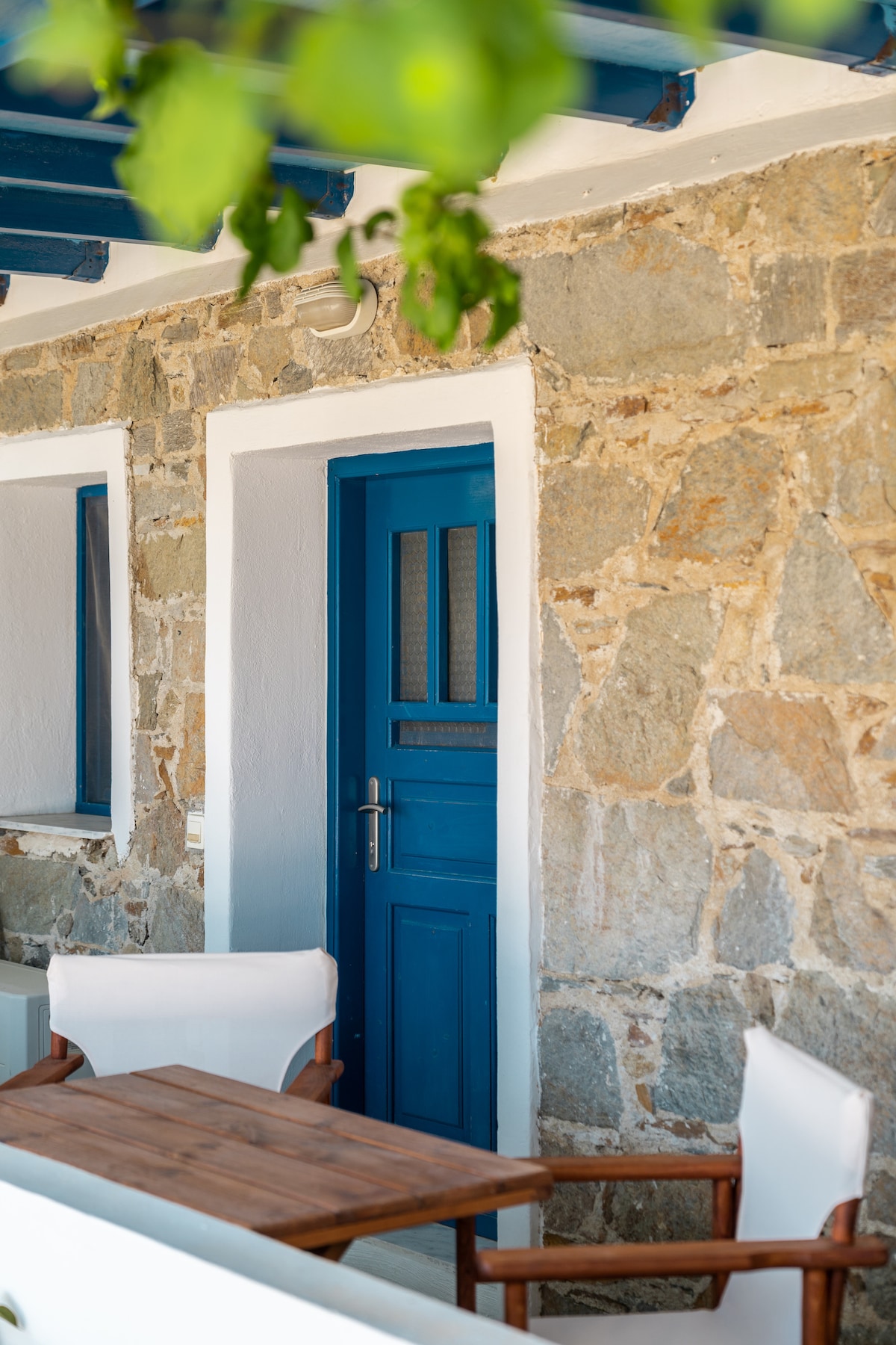 A charming exterior is showcased with a vibrant blue door framed by rustic stone walls. A small wooden table and two chairs are positioned nearby, inviting relaxation. Greenery can be seen overhead, adding a touch of nature to the setting.