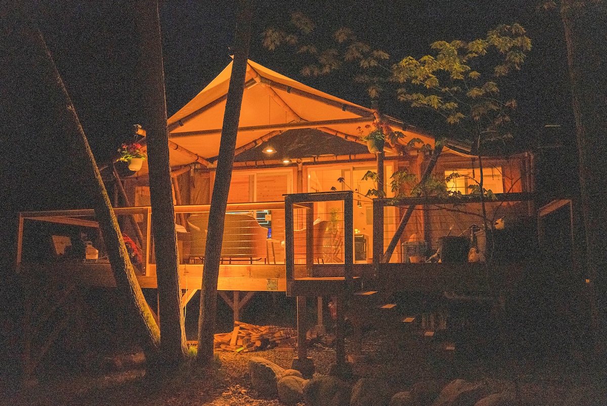 The lodge is illuminated warmly at night, showcasing its elevated wooden deck surrounded by trees. Soft light highlights the canvas structure, creating a serene setting. Outdoor seating is visible on the deck, inviting relaxation in this tranquil atmosphere.