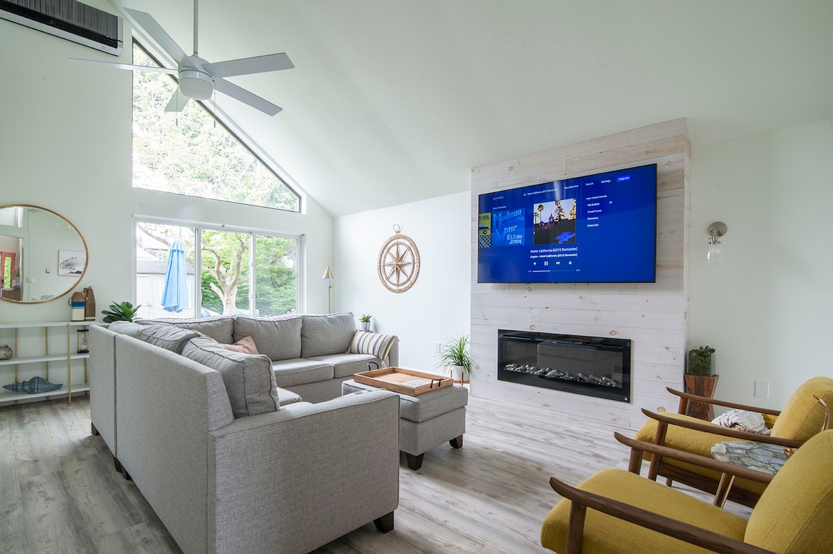 A spacious living area is presented with a sectional sofa and a stylish coffee table. Large windows allow natural light to fill the room. A mounted television is featured above a modern electric fireplace, complemented by two armchairs providing additional seating.