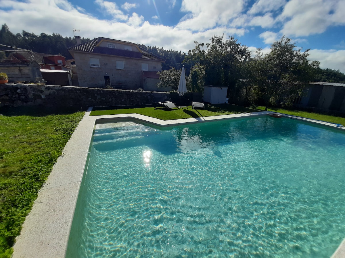 A tranquil outdoor area features a clear, inviting pool surrounded by green grass and sun loungers. The sky above is bright with scattered clouds, and a stone building is visible in the background, enhancing the serene setting.