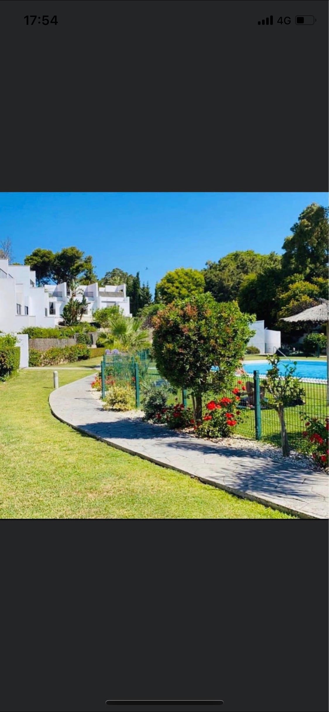 A well-maintained garden area features a winding pathway bordered by shrubs and flowers. A green fence encloses a pool area, with trees and the white buildings visible in the background against a clear blue sky.
