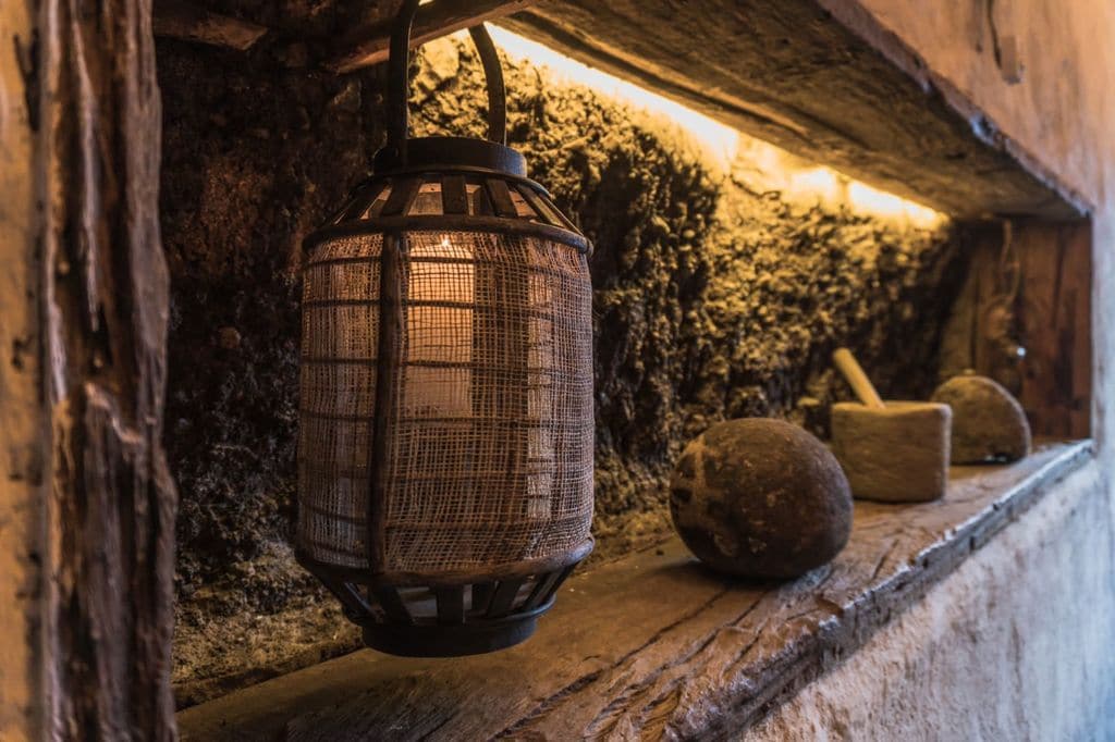 A rustic lantern, crafted with a wire frame, is placed on a wooden shelf next to smooth stones and a bowl. The natural texture of the earthen wall behind is softly illuminated by a warm light fixture above, creating an inviting focal point.