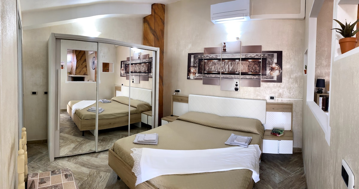 A cozy bedroom features a spacious bed adorned with neutral bedding. Mirrored wardrobe doors reflect the room's light. Wall art adds a decorative touch, and an air conditioning unit is mounted above. Soft lighting enhances the calming atmosphere, while a small plant sits on a shelf.