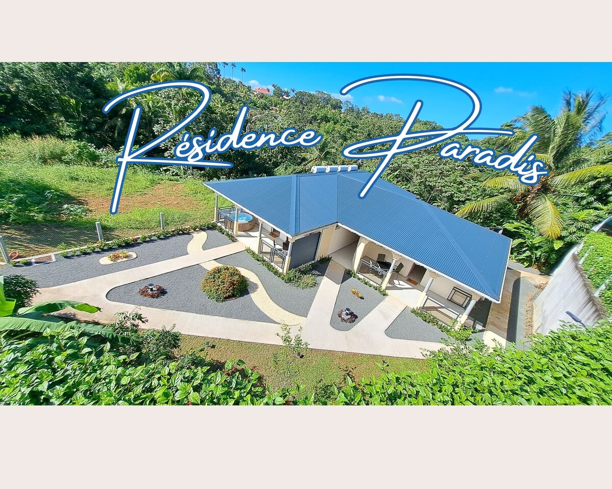 An aerial view of the elegant residence is displayed, showcasing the modern architecture and landscaped gardens. The property features a sloped roof and is surrounded by lush greenery, creating a serene atmosphere. Pathways lead to various outdoor areas, enhancing the tranquil setting.