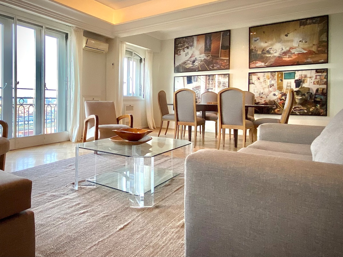 A spacious living area is highlighted by large windows, allowing natural light to fill the room. Comfortable seating options surround a glass coffee table, while a dining area with a wooden table and upholstered chairs is positioned nearby. Art pieces adorn the walls, enhancing the decor.