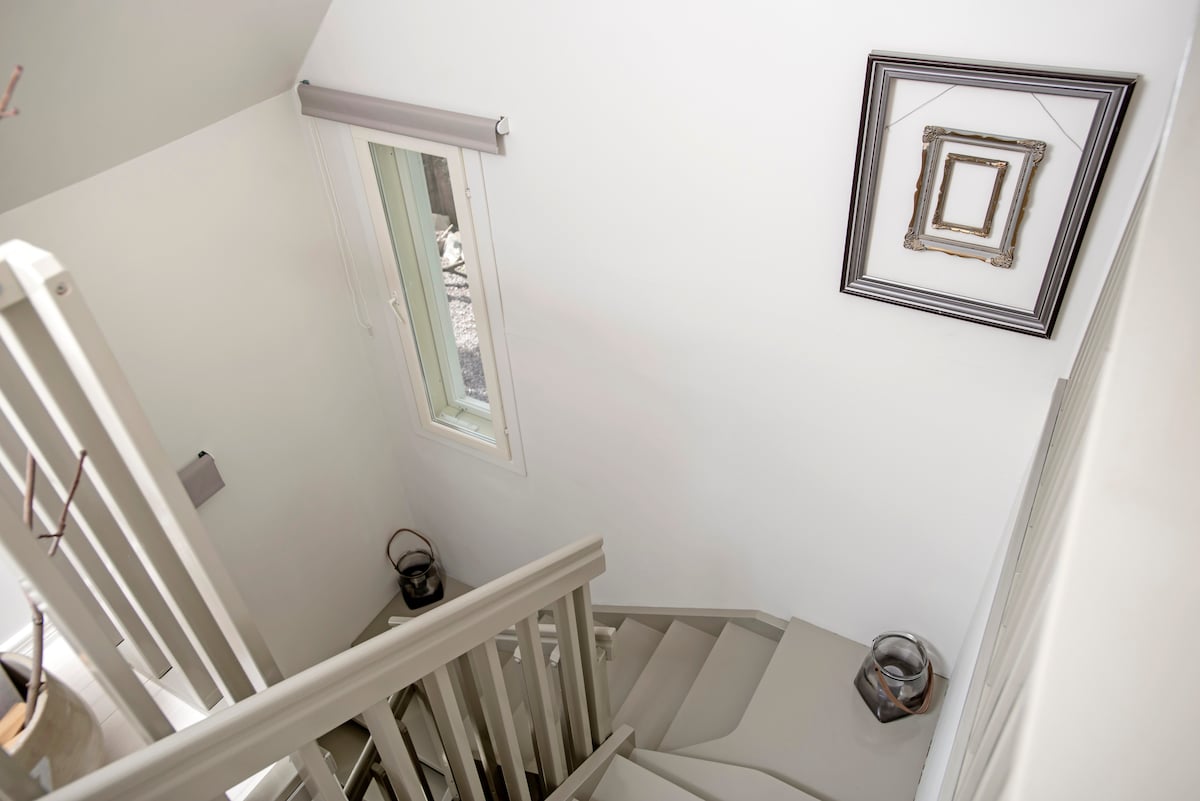 A view of the staircase leading to the upper levels, featuring light-colored walls and steps. A framed artwork hangs beside a window that allows natural light to enter. Decorative elements are placed on either side of the stairs, enhancing the welcoming atmosphere.