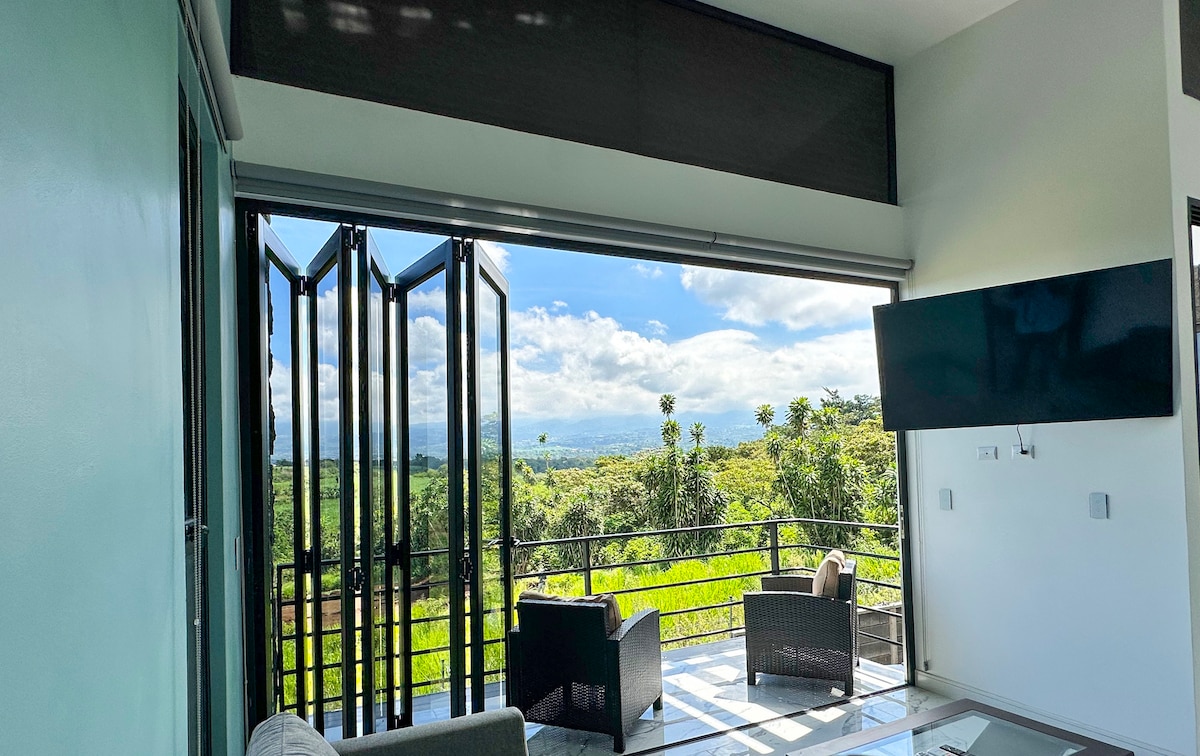 A bright living space is displayed, featuring large glass doors that open to breathtaking mountain views. Two comfortable chairs are positioned in front of the doors, inviting relaxation. Natural light floods the room, highlighting the modern decor and serene atmosphere.