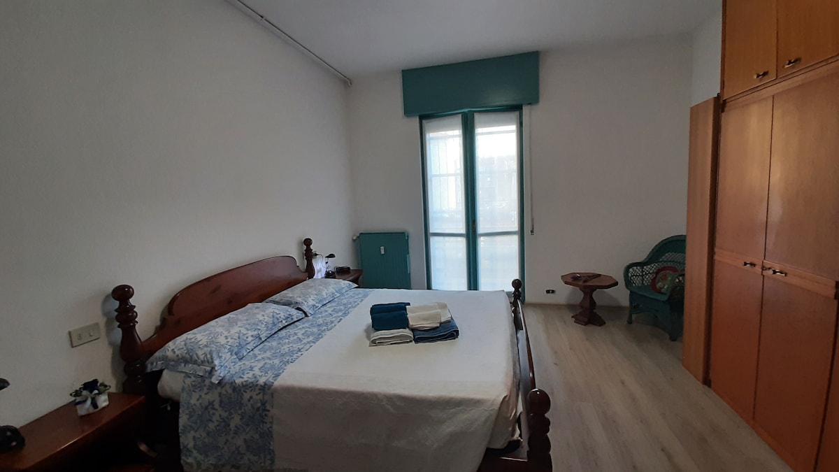 A spacious bedroom features a wooden bed with light bedding and neatly folded towels. Natural light enters through a large window, complemented by a blue decorative chair and wooden furniture. Ample storage is provided by built-in wardrobes along the wall.