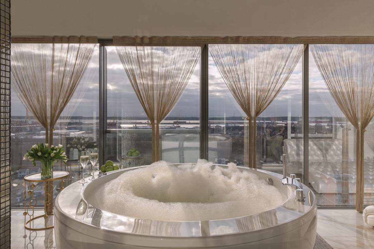 A luxurious jacuzzi is positioned beside large floor-to-ceiling windows, offering scenic views of the surrounding area. Sunlight gently filters through sheer curtains, creating a relaxed atmosphere. Two elegant glasses and a small bouquet sit on a nearby table, enhancing the space for a calming retreat.