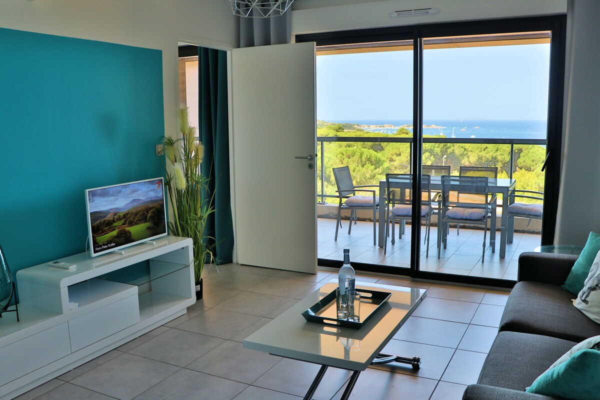 A spacious living area is shown, featuring a flat-screen television and a low coffee table. Large glass doors offer a view of the outdoors, which includes a dining space on a balcony overlooking lush greenery and the sea.