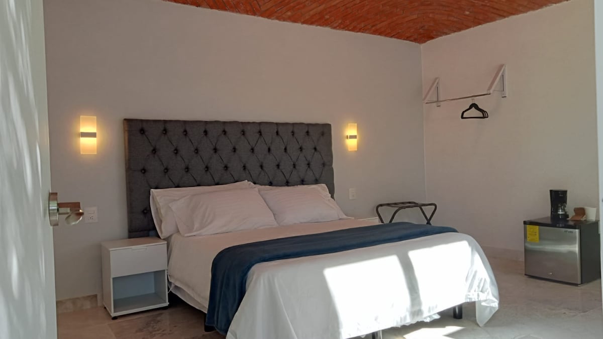 A spacious sleeping area is highlighted by a comfortable bed with a tufted headboard. Soft light fixtures frame the walls, while a small bedside table complements the decor. A clothing rack and a coffee station add functional elements to the room.