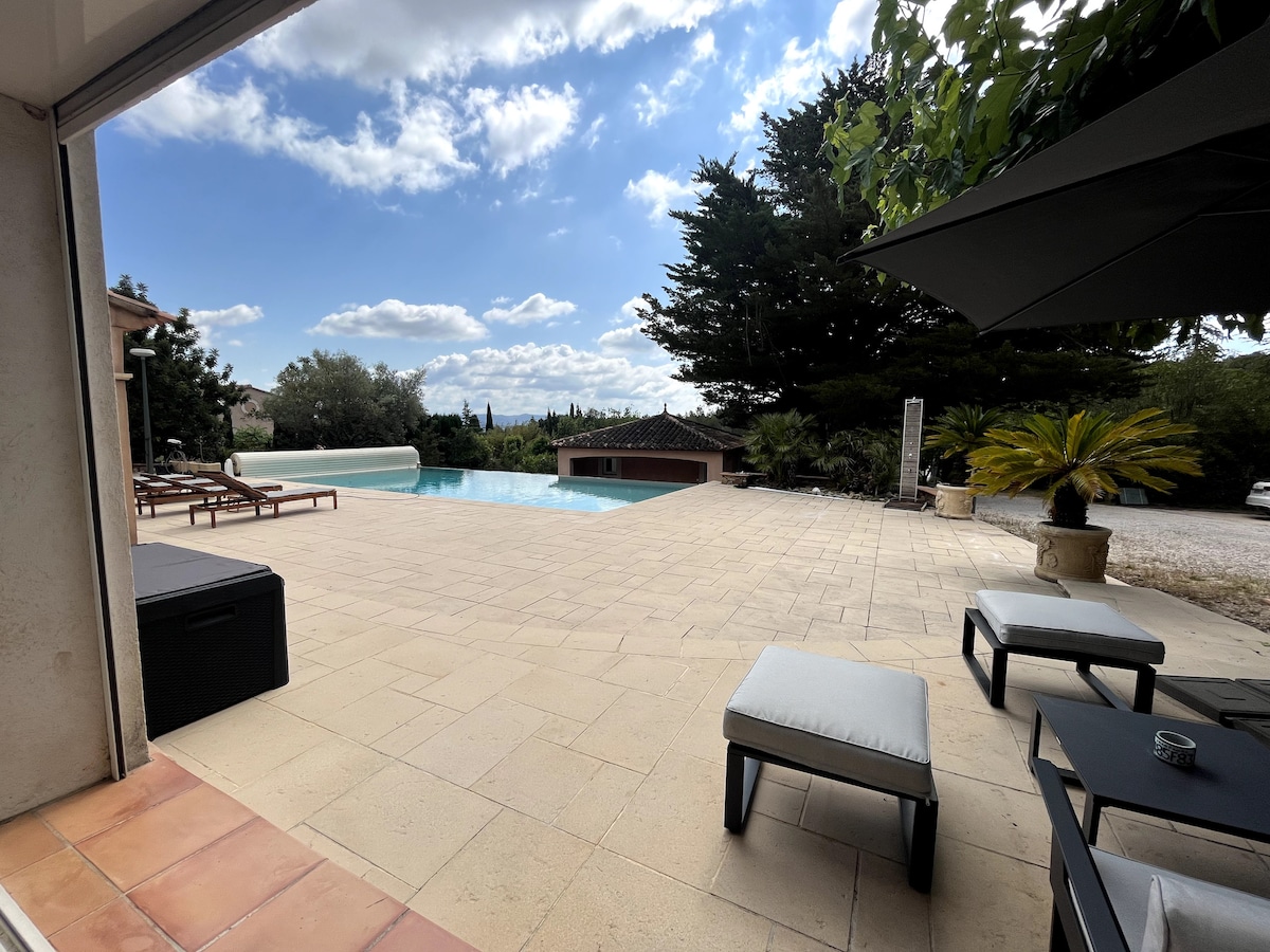 An outdoor space features a spacious terrace with loungers and a seating area. A swim-up pool is visible, surrounded by greenery. The sky displays scattered clouds, and the overall atmosphere is serene and open.