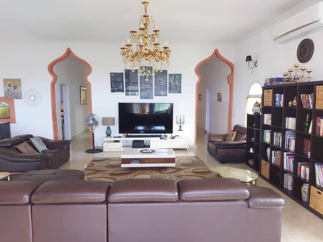 A spacious living room features elegant archways and a decorative chandelier overhead. A large television is mounted on the wall opposite a comfortable seating arrangement, which includes dark leather sofas. A modern coffee table is centered on a patterned rug, while a bookshelf filled with various titles stands nearby.