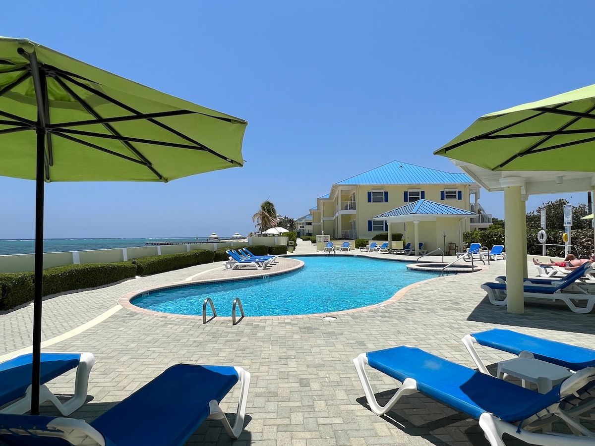 A spacious swimming pool is surrounded by lounge chairs beneath bright green umbrellas. The ocean is visible in the background, while the well-maintained patio features brick pavers. A resort building with multiple windows and a blue roof is situated in the distance.