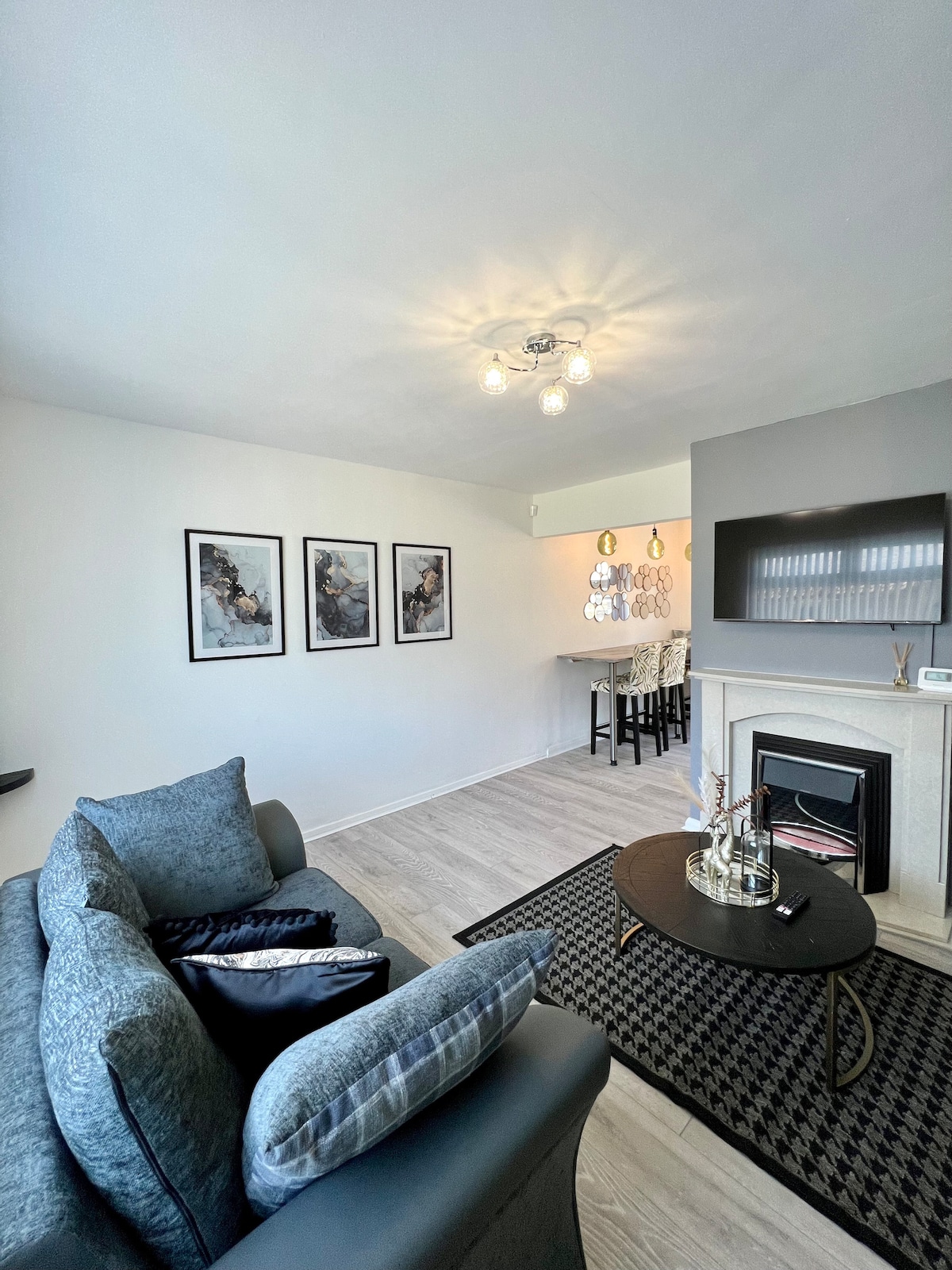 A modern living space is featured with a comfortable gray sofa adorned with patterned cushions. Three framed artworks are displayed on the wall, while a small round table with chairs can be seen in the background. A sleek fireplace adds a touch of warmth to the room.