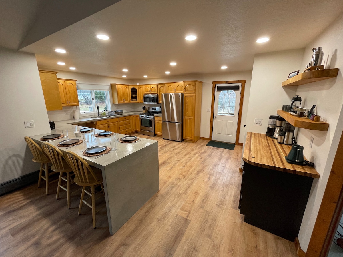 The kitchen features warm wooden cabinetry and modern stainless-steel appliances. A spacious concrete island with seating for four is paired with a dining area. Natural light brightens the room, while a side door provides access to the outdoors.