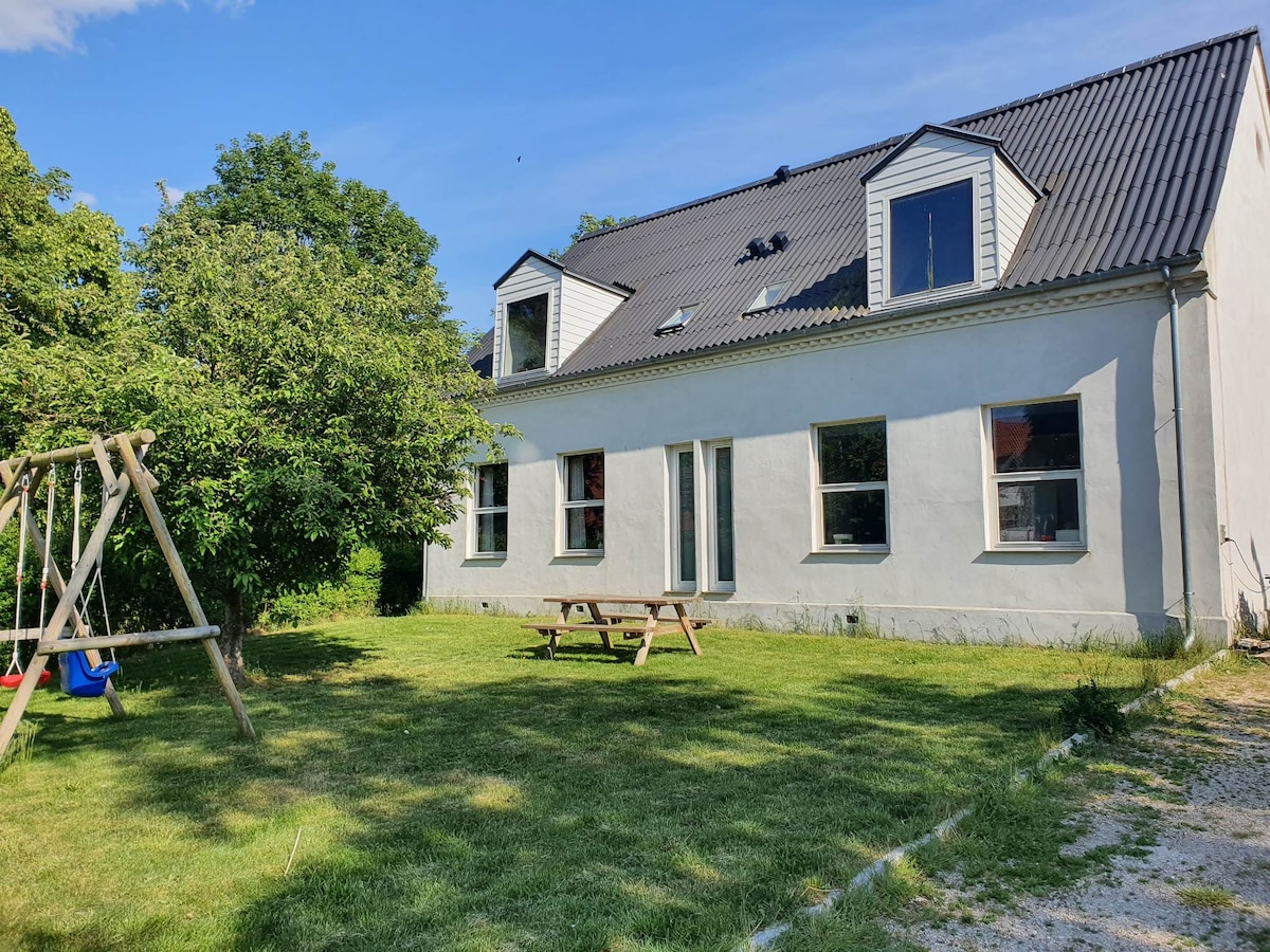 A spacious yard is depicted with a green lawn bordered by trees. A wooden picnic table rests on the grass, accompanied by a swing set featuring two seats. The two-story house, with multiple windows and a sloped roof, provides a bright and inviting exterior.
