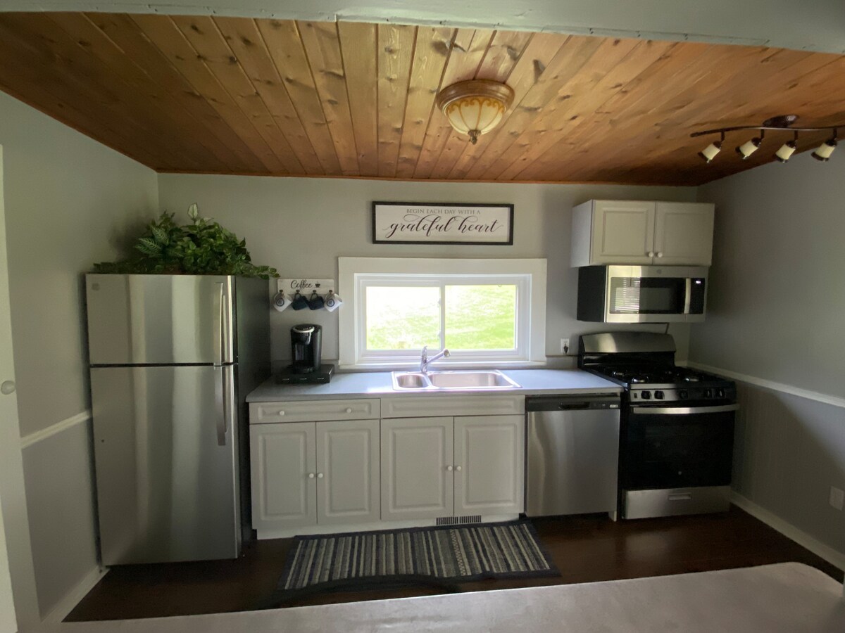 The kitchen area features modern stainless-steel appliances, including a refrigerator and stove. A double sink is positioned under a window that allows natural light. Light-colored cabinets complement the wood-paneled ceiling, while a small coffee maker is available for use.