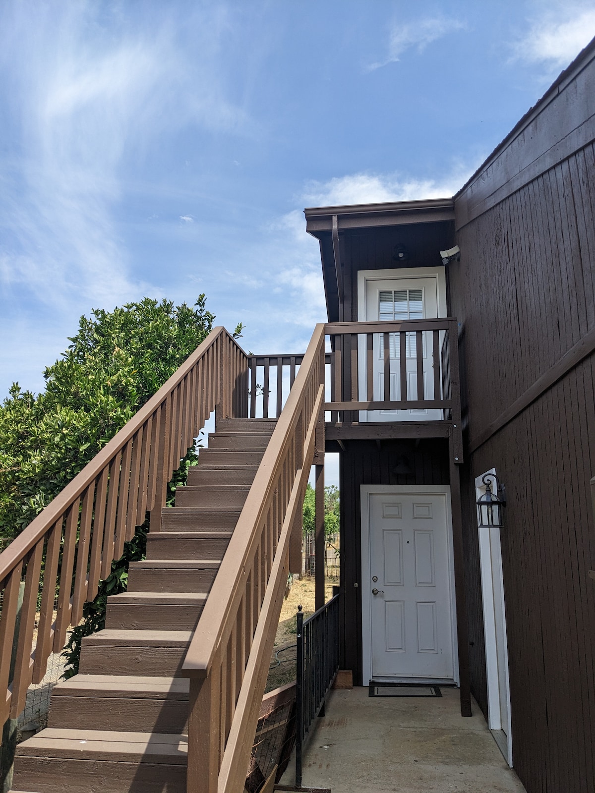 A wooden staircase leads to the entrance of the studio, positioned on the second floor of a brown exterior building. The white door is framed by a lantern and complemented by greenery nearby. The sky above is bright with soft clouds.