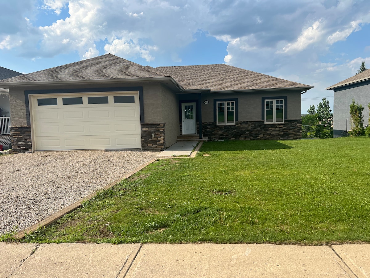 A modern single-story house features a well-maintained front lawn and a gravel driveway leading to a spacious garage. The exterior showcases a combination of light-colored walls and stone accents, under a gently sloping roof. Clear skies provide a bright backdrop.