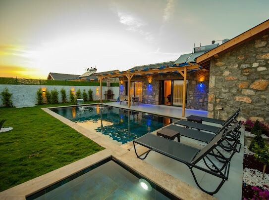 The outdoor area features a stone house beside a rectangular pool, surrounded by a lawn. Sun loungers are arranged around the pool, and warm lighting accentuates the structure in the evening glow. A harmonious blend of natural and architectural elements is presented.