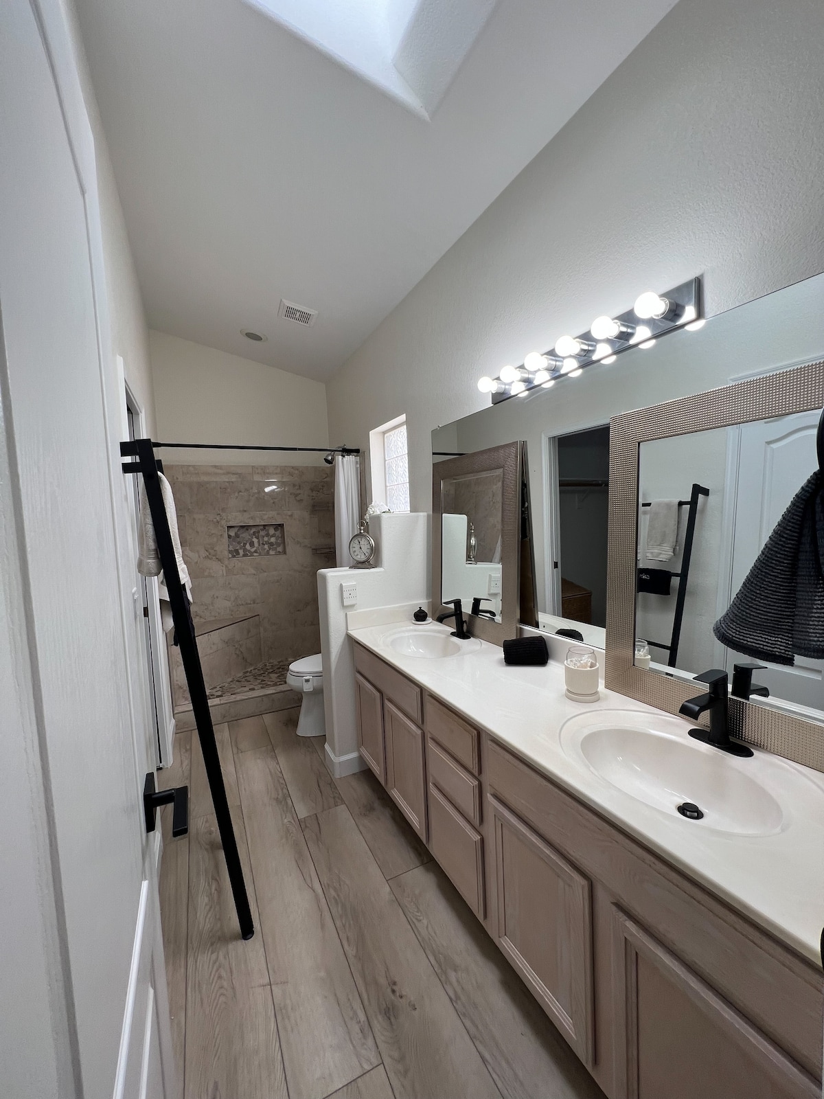 A well-appointed bathroom features dual sinks set in light-toned cabinetry. A spacious shower is enclosed with glass, showcasing intricate tile work. Natural light filters through a skylight, illuminating the neutral color palette and enhancing the clean lines of the design.
