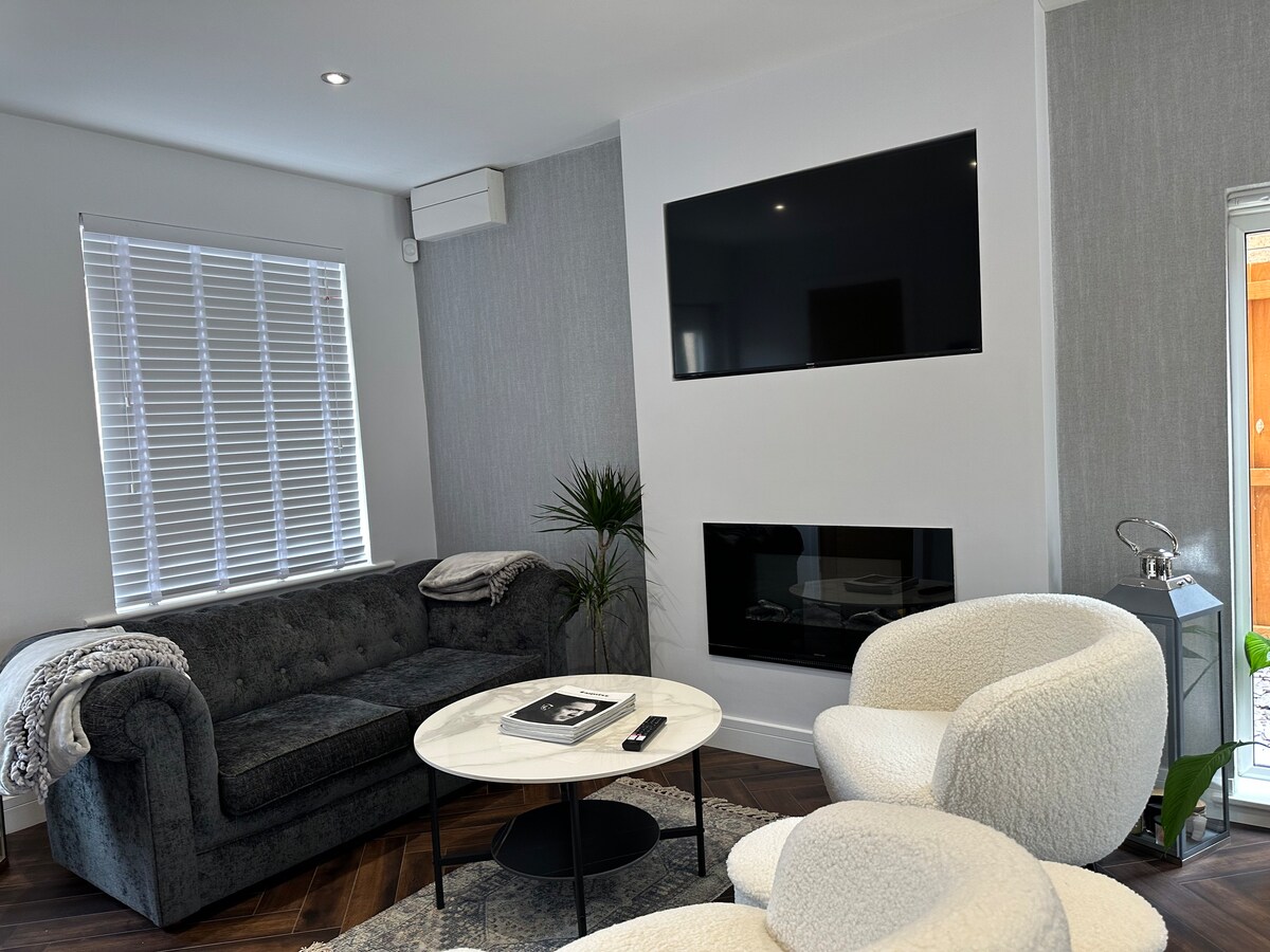 A modern living area features a plush sofa and stylish armchairs, set around a round coffee table. A sleek flat-screen TV is mounted on the wall above a contemporary fireplace. Natural light filters through a window adorned with white blinds.