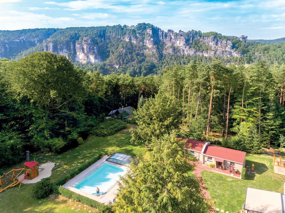 A scenic view of the resort's outdoor area, featuring a swimming pool surrounded by lush greenery. The backdrop consists of rocky sandstone formations, with tall trees providing a natural border. An inviting atmosphere is created by the open space, suitable for relaxation and leisure activities.
