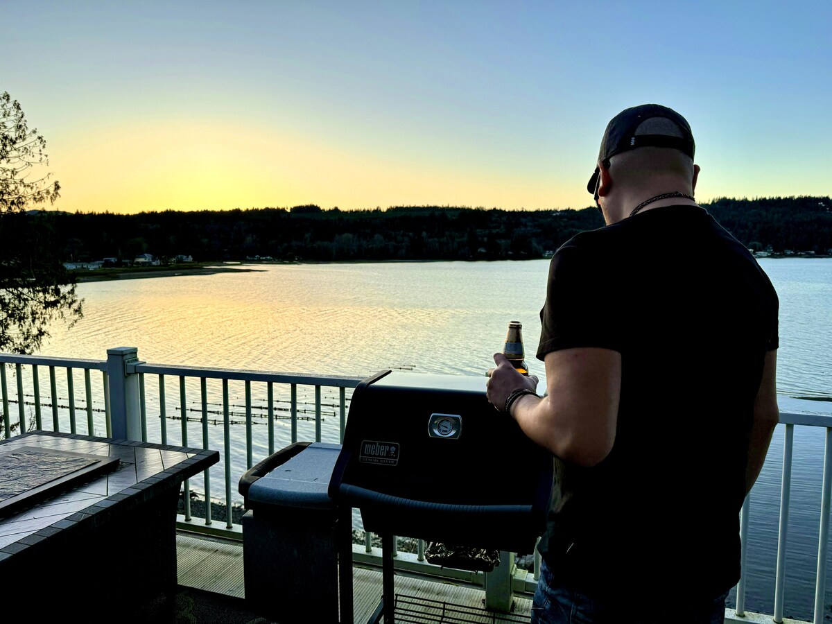 A person stands by a gas grill on a private waterfront patio, overlooking a serene bay at sunset. The calm water reflects hues of orange and blue, while trees frame the distant shoreline. A beverage is held in one hand, enhancing the peaceful outdoor atmosphere.