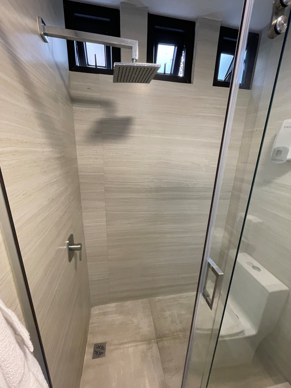 A contemporary shower area features beige tiled walls and a glass enclosure. A modern showerhead is positioned overhead, with natural light filtered through small windows above. A minimalist toilet is visible within the space, and a towel is hung on a nearby hook.