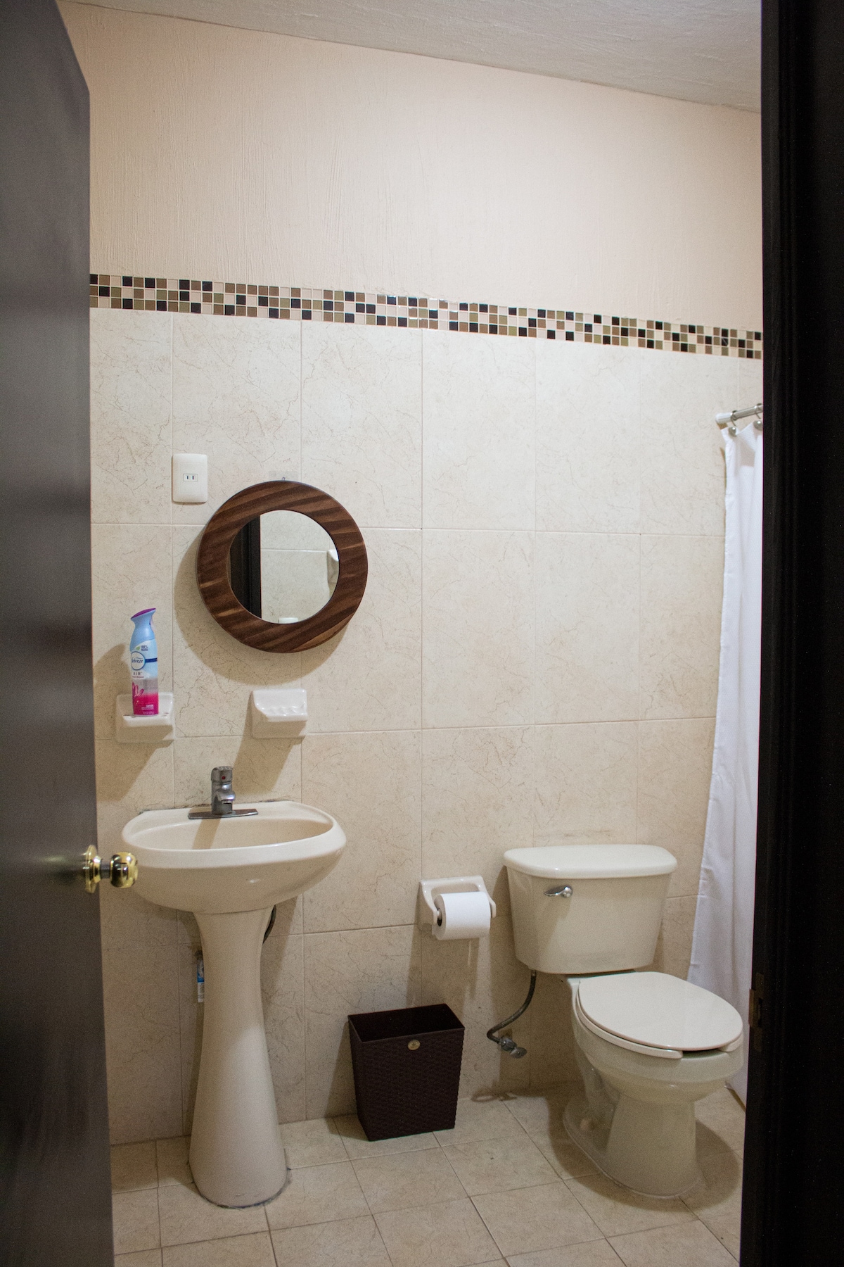 A compact bathroom features light-colored tiles and a circular mirror above a pedestal sink. A toilet is positioned beside a waste bin. A shower area is enclosed with a white curtain, while a decorative strip of small tiles runs along the upper wall.