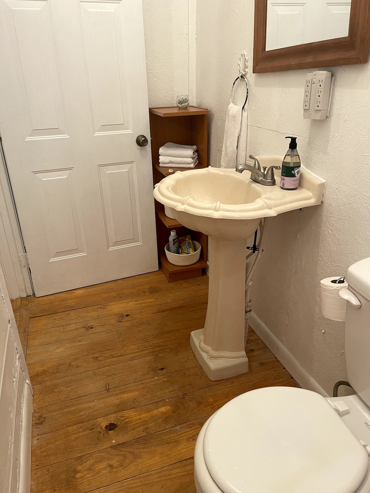 A full-size bathroom features a pedestal sink with a soap dispenser and toiletries. Wooden flooring adds warmth to the space, while a toilet is positioned nearby. A cabinet is visible, stocked with neatly folded towels, contributing to the functional design.