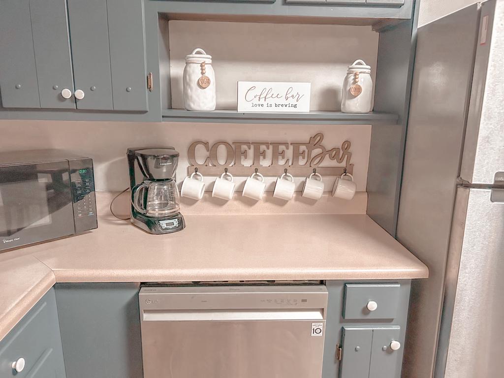 A functional kitchen corner features a coffee bar setup with six hanging coffee mugs. A coffee maker sits beside a microwave, and a sign reading 'Coffee Bar' is displayed on the shelf above. The countertops are clean and uncluttered, enhancing the space's utility.