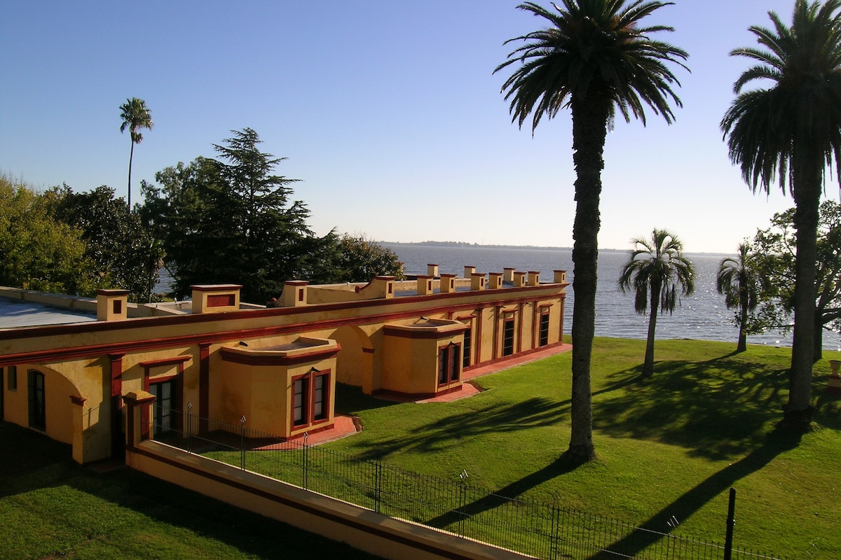 A low, yellow building with multiple windows is set against a serene water view. Large palm trees and green lawns create a spacious outdoor area, enhancing the tranquil environment. The clear sky and gentle waters contribute to a calm and relaxing atmosphere.