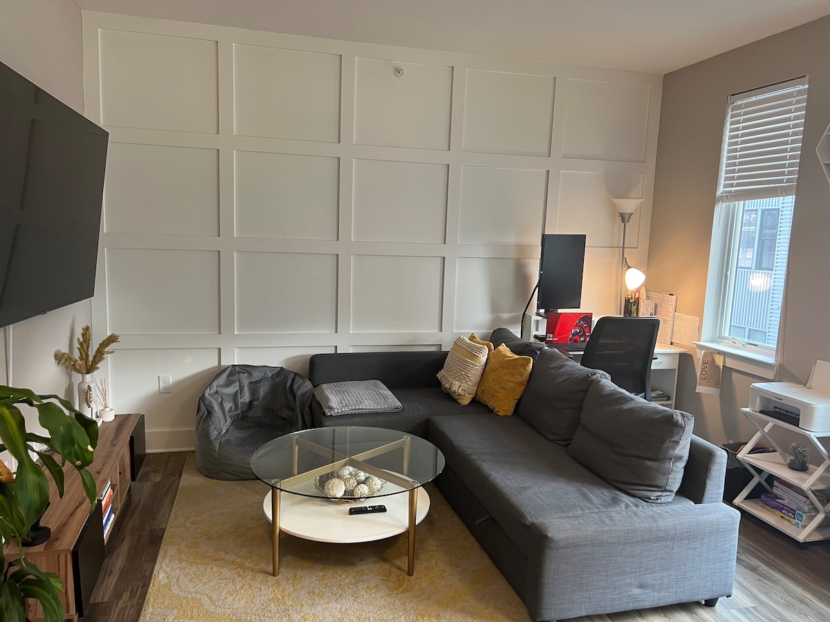 A modern living area features a sectional couch in gray, complemented by a round coffee table with decorative items. A bean bag is situated nearby, and a workspace with a desk and chair is illuminated by natural light from a window.