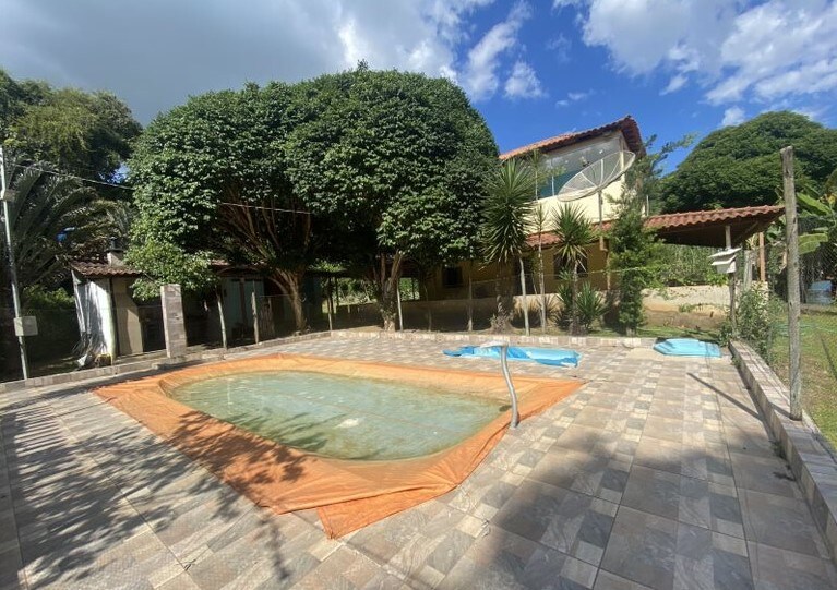 A spacious outdoor area features a large pool, partially covered with an orange tarp, surrounded by a tiled deck. Lush greenery, including tall trees and shrubs, adds a natural element to the setting. Smaller pools are visible nearby, providing additional options for relaxation.