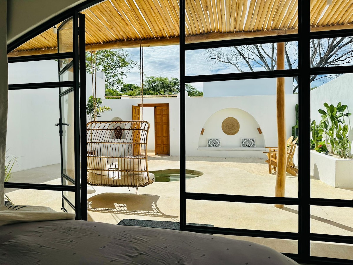 A view from inside the villa showcases a private garden with a swimming pool. A hanging chair and lounge seating are positioned around the pool area, with natural light flooding in through large glass doors. Lush greenery and a rustic wooden door add to the serene ambiance.