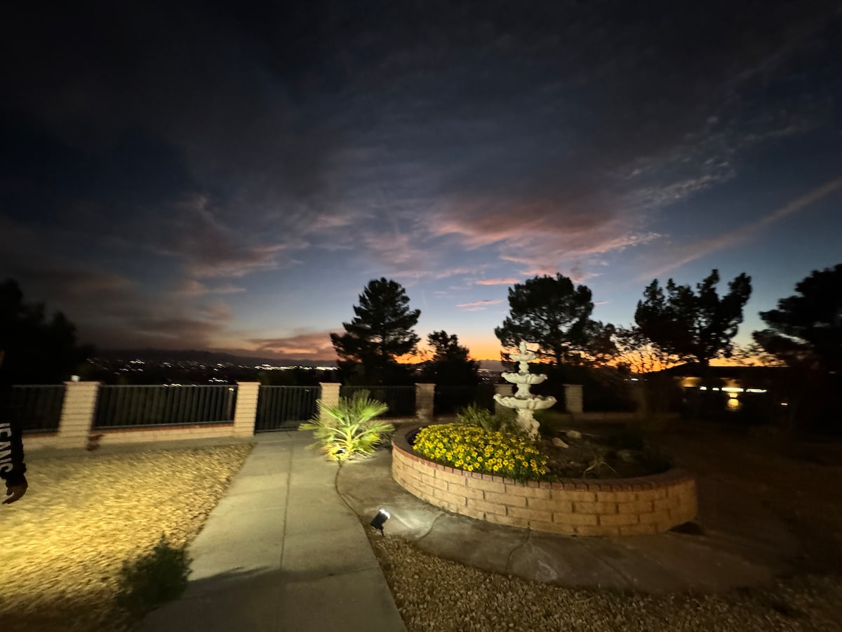 The image captures a tranquil outdoor space at dusk, featuring a well-maintained pathway leading toward a garden adorned with yellow flowers and a stone fountain. Silhouettes of trees line the background, while a subtle glow from nearby lights enhances the serene atmosphere.