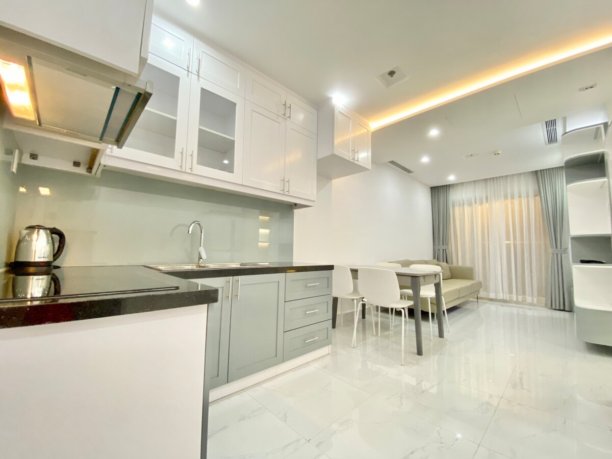 A modern kitchen is complemented by sleek cabinetry and high-efficiency appliances. A dining table with white chairs is positioned near a cozy living area, featuring a light-colored sofa. Natural light filters through sheer curtains, illuminating the polished tile flooring.
