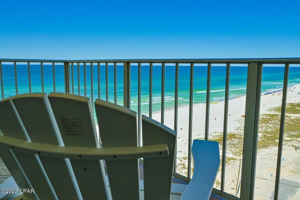 A relaxing balcony chair is positioned on a railing, offering a view of the sandy beach and vibrant blue ocean. Gentle waves can be seen lapping the shore, and the clear sky enhances the coastal ambiance.