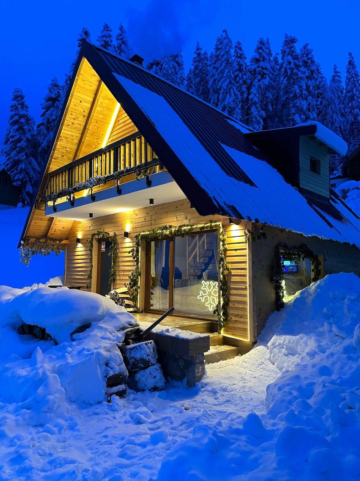 A charming villa is surrounded by snow-covered terrain, showcasing a sloped roof and wooden exterior illuminated by warm lights. A large front deck features decorative elements, while expansive glass doors reflect the cozy interior. Tall trees dusted with snow provide a serene backdrop.