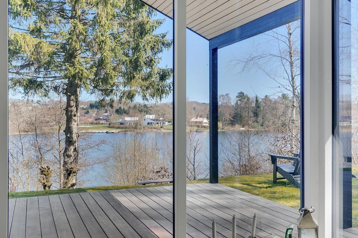A covered outdoor space features a wooden deck overlooking a calm lake surrounded by trees. A large glass panel provides unobstructed views of the serene water and distant homes, while a single chair is visible on the deck, inviting relaxation.