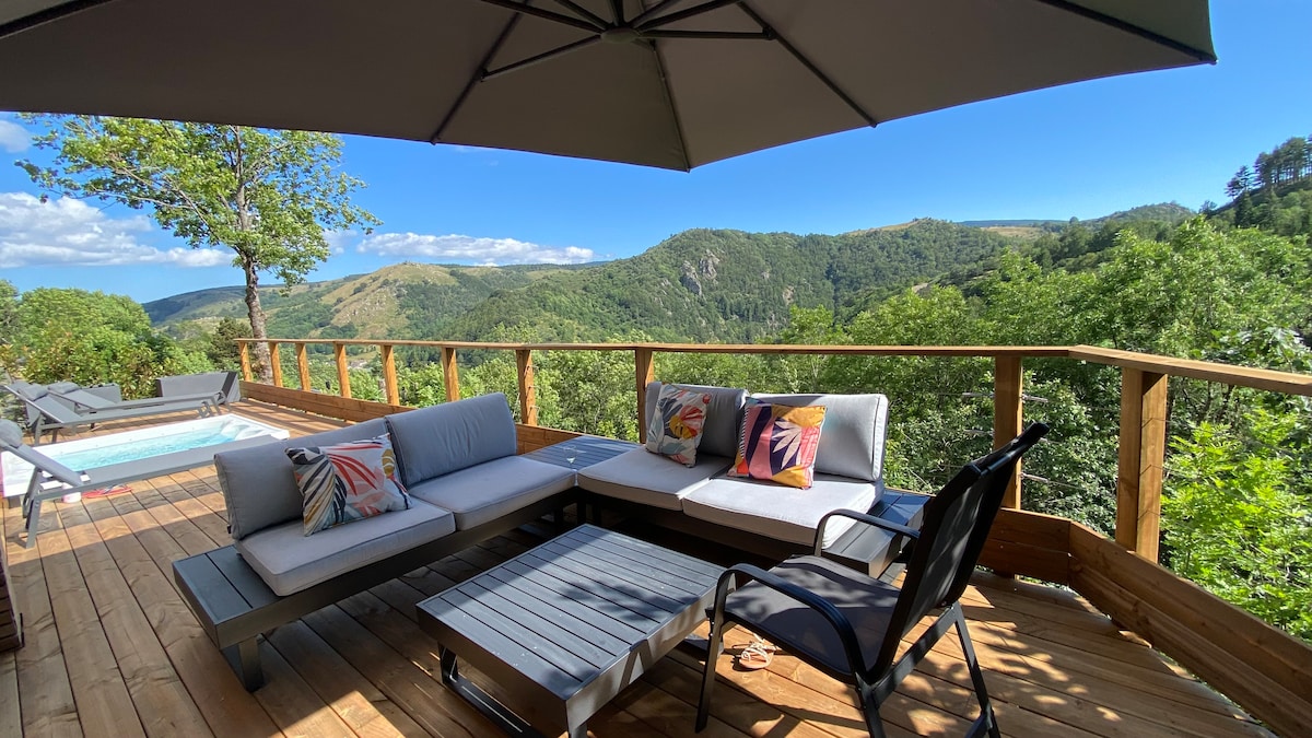 A wooden terrace features a cozy seating area with grey cushions and colorful pillows, providing an inviting spot to enjoy the mountain views. A parasol offers shade, while a private jacuzzi adds a relaxing touch to the outdoor experience.