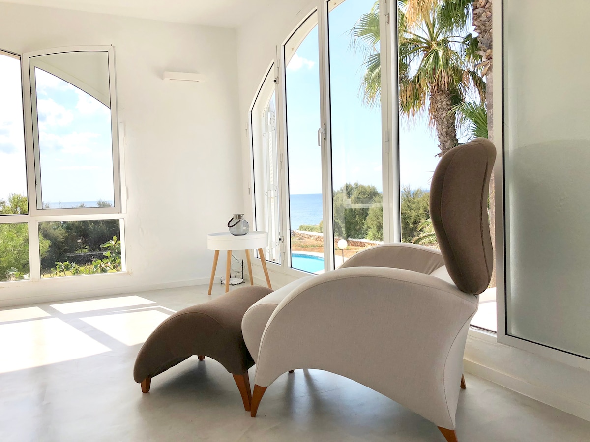 A modern seating area is featured, showcasing a stylish armchair with a chaise lounge design. Large windows provide views of the outdoor scenery, including palm trees and the distant sea. Natural light enhances the tranquil atmosphere.