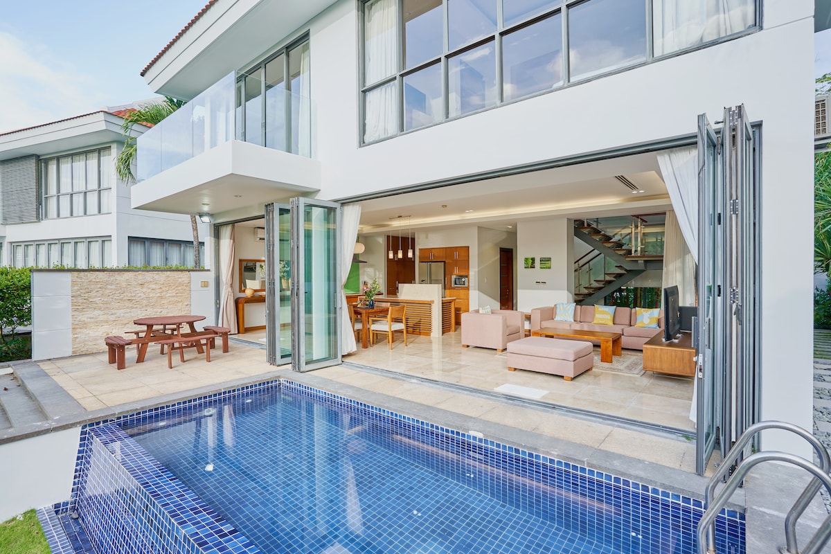 The outdoor area features a private pool surrounded by a spacious terrace. Large glass doors connect the living space to the poolside, allowing for natural light. A dining table is visible, alongside comfortable seating arranged indoors, promoting a welcoming environment.