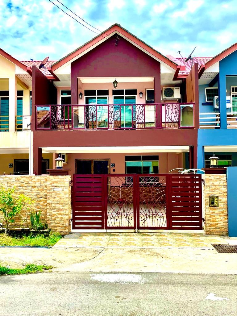 A vibrant two-story house features a combination of purple and light-colored facades, framed by decorative red gates. The entrance showcases a balcony with multiple windows, while stone accents and greenery enhance the welcoming appearance of the property.